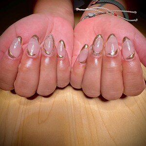 May include: Close-up of hands with nude-coloured oval-shaped nails with a gold accent around the edge of each nail.