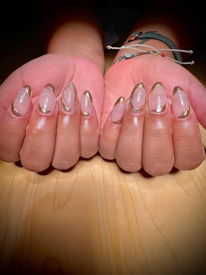 May include: Close-up of hands with nude-coloured oval-shaped nails with a gold accent around the edge of each nail.