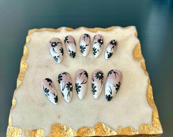 以下が含まれることがあります： 白とピンクのグラデーションデザインと黒い花のアクセントが付いた、10本の付け爪のセットです。