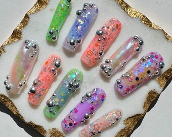 May include: Ten colourful press-on nails with glitter and silver accents. The nails are arranged on a white marble surface with a gold border.