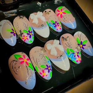 May include: A set of ten false fingernails with a light blue base and floral designs. Each nail has a different design featuring flowers, leaves, and glitter. Some nails have a white bow with a pearl accent.