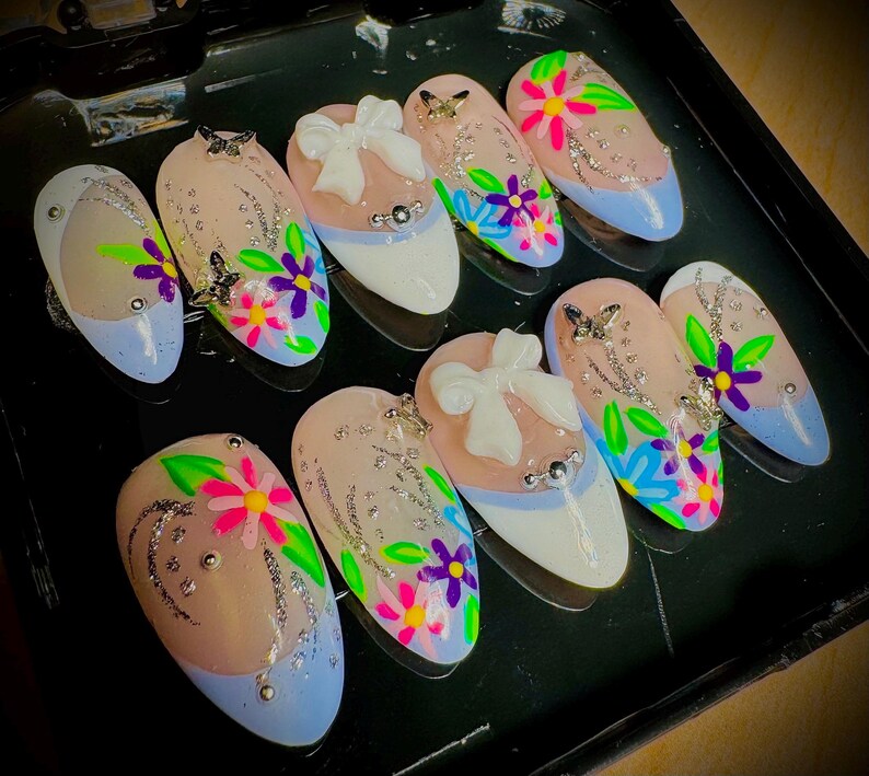 May include: A set of ten false fingernails with a light blue base and floral designs. Each nail has a different design featuring flowers, leaves, and glitter. Some nails have a white bow with a pearl accent.