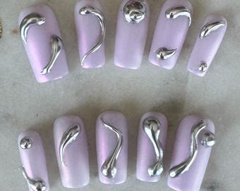 May include: Ten light purple artificial nails with silver metallic designs. The designs are abstract and resemble drips or swirls.