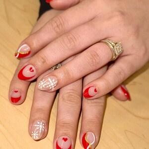 May include: Close-up of hands with red and white nail polish. The nails are decorated with hearts, snowflakes, and gold accents.