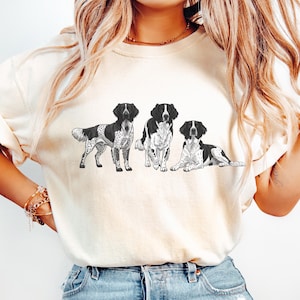 May include: Cream-colored t-shirt featuring a black and white illustration of three dogs. The dogs are depicted in different poses: standing, sitting, and lying down. The graphic is centered on the shirt.