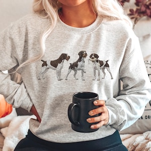 Pode incluir: Camisola cinza claro com três cães da raça Braco Alemão em fila. Os cães são castanhos e brancos, um deles segura um objeto na boca. Uma caneca preta é segurada em primeiro plano.