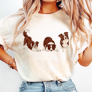 May include: Cream-colored t-shirt featuring a playful illustration of three Border Collies in different poses. The dogs are depicted in brown and white, with one stretching, one lying down, and one standing.