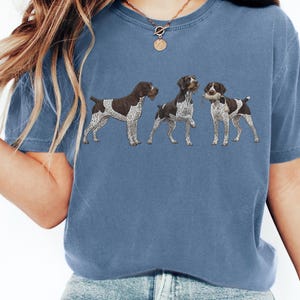May include: A blue t-shirt featuring three German Shorthaired Pointers in a row. The dogs are brown and white, with detailed illustrations. The shirt is a solid blue color, and the overall style is casual.