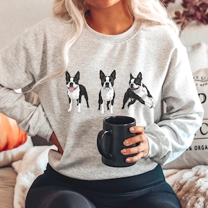May include: A light gray sweatshirt featuring three Boston Terrier illustrations in black and white. The sweatshirt is a crew neck style and is made of a soft material. The person is holding a black mug.