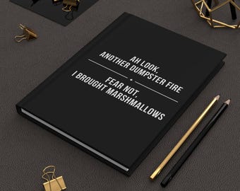 May include: Black journal with the text "AH LOOK, ANOTHER DUMPSTER FIRE, FEAR NOT, I BROUGHT MARSHMALLOWS" in white. Gold and black pencils and gold binder clips are also on the dark gray surface.
