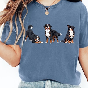 Könnte beinhalten: Blaues T-Shirt mit drei Berner Sennenhunden in verschiedenen Posen. Die Hunde sind schwarz, braun und weiß, mit detaillierten Illustrationen. Das T-Shirt hat einen lässigen, bequemen Stil.