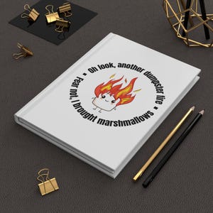 May include: White journal with the text "Oh look, another dumpster fire. Fear not, I brought marshmallows." The journal features a cartoon marshmallow on fire. Gold and black pencils and binder clips are also visible.