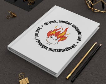 May include: White journal with the text "Oh look, another dumpster fire. Fear not, I brought marshmallows." The journal features a cartoon marshmallow on fire. Gold and black pencils and binder clips are also visible.