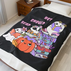May include: Black throw blanket with a whimsical design featuring cartoon ghosts, a pumpkin, and a skull reading books. The text "Hot Ghouls Read" is displayed in pink. The blanket is on a bed with a white pillow.