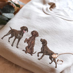 Puede incluir: Sudadera color crema con tres ilustraciones de perros de raza Braco Alemán en varias poses. Un perro está de pie, otro sentado y el tercero se levanta. Los perros son marrones con marcas blancas.