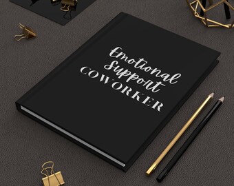 May include: A black journal with the words "Emotional Support COWORKER" in white script. Gold and black pencils, along with gold binder clips, are arranged around the journal on a dark gray surface. A decorative gold geometric object is in the background.
