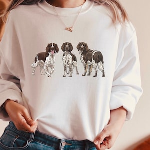 Könnte beinhalten: Weißes Sweatshirt mit einer Grafik von drei English Springer Spaniels. Die Hunde sind in Braun und Weiß dargestellt, wobei die Zunge eines Hundes sichtbar ist. Das Sweatshirt hat einen Rundhalsausschnitt und lange Ärmel.