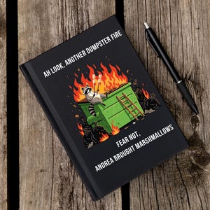May include: Black journal with the text "AH LOOK, ANOTHER DUMPSTER FIRE" and "FEAR NOT, ANDREA BROUGHT MARSHMALLOWS." The cover features a raccoon in a dumpster on fire. A black pen rests on a wooden surface.
