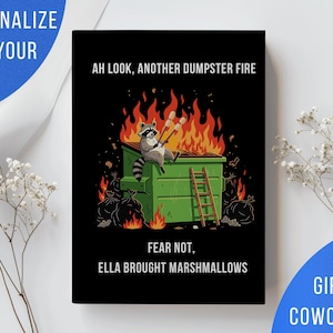 May include: A black rectangular print featuring a cartoon raccoon sitting on a green dumpster engulfed in orange and red flames. The raccoon holds two marshmallows on sticks. Text reads: AH LOOK, ANOTHER DUMPSTER FIRE FEAR NOT, ELLA BROUGHT MARSHMALLOWS.