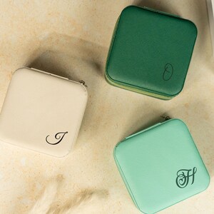 May include: Three square, zippered jewelry cases in cream, green, and mint green. Each case has a different initial in black script. The cream case has the letter "J", the green case has the letter "O", and the mint green case has the letter "H".