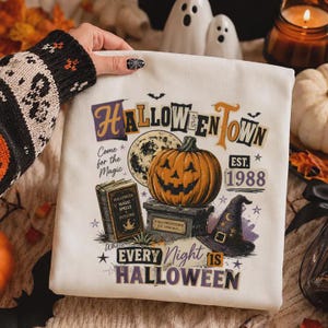 May include: A white sweatshirt featuring a graphic print with the text HALLOWEENTOWN EST. 1988, Come for the Magic, and EVERY NIGHT IS HALLOWEEN. The design includes a jack-o'-lantern, a full moon, a spellbook, and a witch hat in orange and purple.
