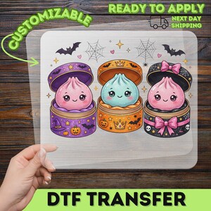 May include: A DTF transfer sheet featuring three cartoon dumplings in round containers. The left is purple with pumpkins and bats, the center is orange with crowns, and the right is black with pink bows and skulls. Bats and spiderwebs decorate the top.
