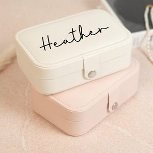 May include: Two rectangular jewelry boxes, one cream-colored and one pink, stacked on top of each other. The top box has the name "Heather" in black script. Both boxes have a silver clasp and are on a light pink surface.