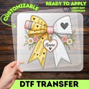 May include: A decorative bow transfer featuring a yellow side with pencil and heart patterns and a white side with lined paper texture. A pink center knot holds a heart-shaped charm with the text Name and a small heart. Floral accents include pink roses and white daisies.