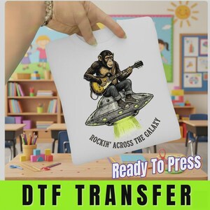 May include: A white t-shirt featuring a graphic of a chimpanzee playing an electric guitar while sitting on a flying saucer. Below the illustration, the text reads Rockin' Across The Galaxy in black lettering. The design is centered on the fabric.