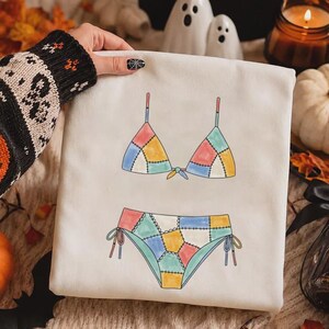 May include: A white sweatshirt featuring a graphic print of a two-piece bikini with a patchwork design. The bikini top and bottom are composed of multi-colored segments in shades of yellow, pink, blue, and green, outlined with black stitching.