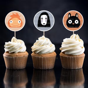 以下が含まれることがあります： 白いフロスティングが施された3つのカップケーキ。それぞれ丸いキャラクターのトッパーが付いています。トッパーは、オレンジ色、黒色、白いマスクのキャラクターの漫画のキャラクターが特徴です。カップケーキは反射する黒い表面にあります。