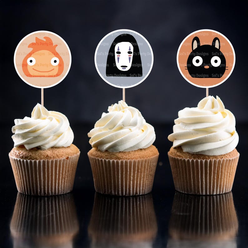 May include: Three cupcakes with white frosting, each topped with a circular character topper. The toppers feature cartoon characters in orange, black, and a character with a white mask. The cupcakes sit on a reflective black surface.