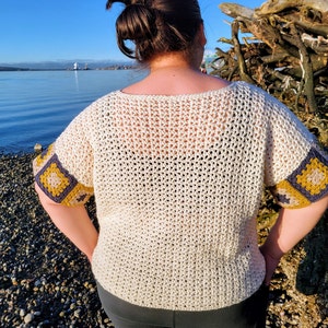 May include: A white crochet top with a loose fit and short sleeves. The sleeves have a crocheted border with yellow and blue squares. The top is worn by a person standing on a beach.