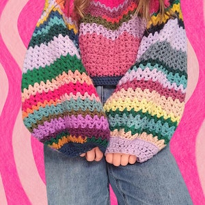 Puede incluir: Un suéter de crochet a rayas de colores con mangas largas y un corte corto. El suéter está hecho con una variedad de colores, incluyendo rosa, morado, verde, azul y amarillo.
