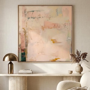 Puede incluir: Interiorismo de una habitación con una gran pintura abstracta en un marco dorado, con suaves tonos rosas y beige. Una lámpara dorada, una consola de mármol y un sillón crema con una manta beige también son visibles.