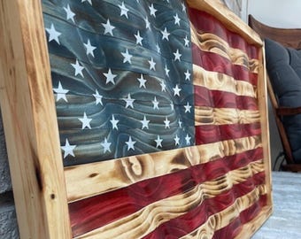 May include: A wooden American flag with a wavy design. The flag features red and white stripes, a blue field with white stars, and a natural wood frame. The wood has a distressed, textured appearance. The frame is made of natural wood.