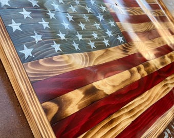Può includere: Un'opera d'arte da parete in legno con un design ondulato della bandiera americana. La bandiera è costruita con assi orizzontali con una finitura in legno bruciato, che mostra strisce alternate rosse e color legno naturale e un cantone blu con stelle dipinte in bianco.