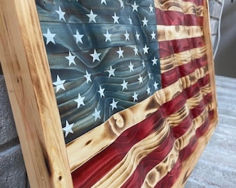 May include: A rectangular wooden wall art piece featuring a carved, wavy American flag design. The flag displays a blue canton with white stars and alternating red and natural wood stripes. The wood has a scorched finish, framed in light wood.