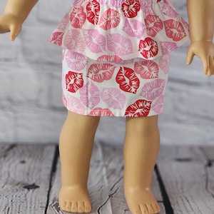 May include: A doll wearing a pink and white dress with a red lip print. The doll has blonde hair and green eyes.