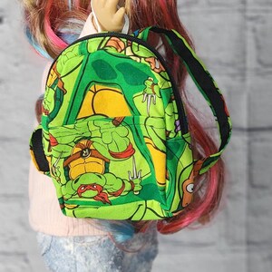 May include: A green backpack featuring a Teenage Mutant Ninja Turtles design. The backpack has black straps and a small handle. The doll is wearing light blue jeans and a pink shirt. The doll has colorful hair.