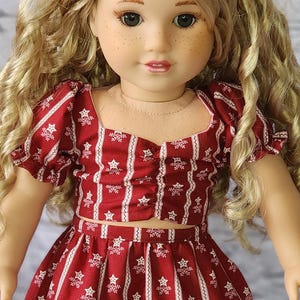 May include: A doll wearing a red and white patterned outfit, including a crop top, tiered skirt, and headband. The fabric features vertical stripes with a floral design. The doll has blonde curly hair and is posed against a light brick wall.