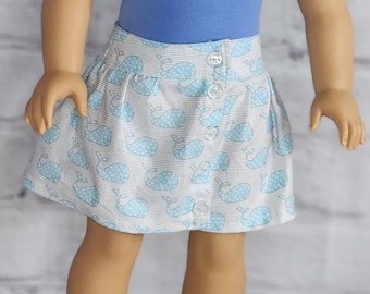 Puede incluir: Una muñeca con un top de tubo azul y una falda abotonada. La falda es blanca con un estampado de ballenas azul claro. La muñeca tiene el pelo largo rubio y ojos verdes. La muñeca está de pie frente a una pared de ladrillos blancos.