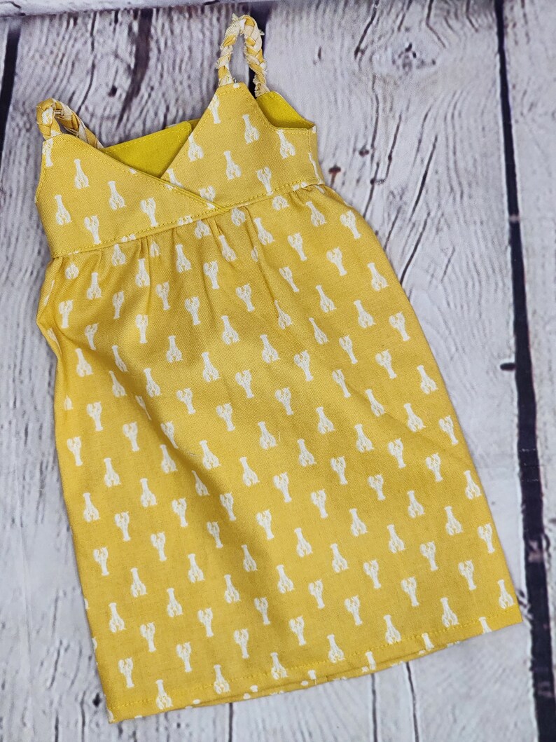 May include: A yellow sundress with white lobster print. The dress features a V-neckline, braided straps, and a gathered waist. The fabric has a soft texture, and the overall design is casual and summery.