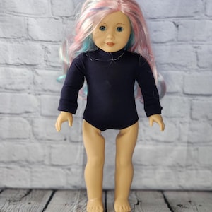 Puede incluir: Una muñeca con ojos azules y pelo largo multicolor en tonos rosa, azul y blanco. La muñeca lleva un maillot de manga larga azul marino. La muñeca está de pie sobre una superficie de madera frente a una pared de ladrillos blancos.