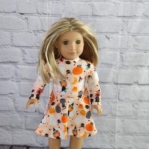 May include: A doll wearing a long-sleeved dress with a Halloween-themed print. The dress features a light peach background with orange pumpkins, black cats, ghosts, and witch figures. The doll has blonde hair and green eyes.