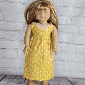 May include: A doll wearing a yellow dress with a white lobster pattern. The dress has a wrap-style bodice and spaghetti straps. The doll has long blonde hair and is standing in front of a white brick wall.
