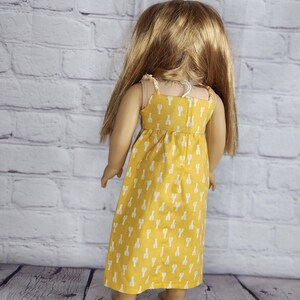 May include: A doll wearing a yellow sundress with white lobster print. The dress has thin straps and a gathered waist. The doll has long blonde hair and is standing in front of a white brick wall.