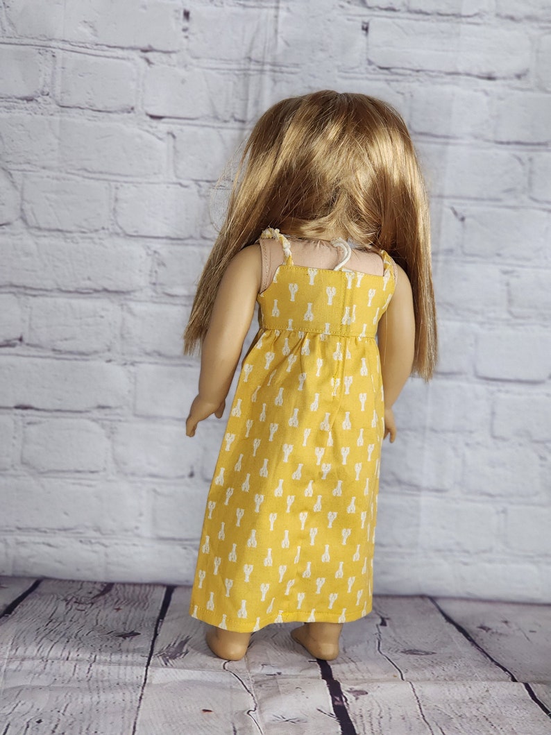 May include: A doll wearing a yellow sundress with white lobster print. The dress has thin straps and a gathered waist. The doll has long blonde hair and is standing in front of a white brick wall.