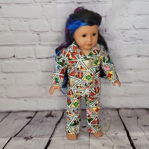 Puede incluir: Una muñeca con un pijama azul claro con un colorido estampado geométrico. El patrón incluye formas triangulares, osos de peluche y las letras "GS". La muñeca tiene el pelo azul y morado.