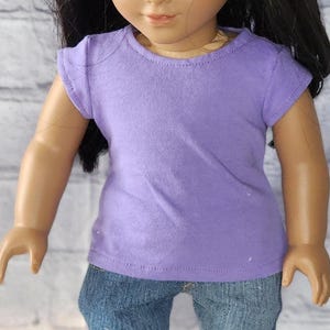 Peut inclure: Un t-shirt lavande à manches courtes et col rond. Le t-shirt est associé à un jean en denim bleu. La tenue est présentée sur une poupée.